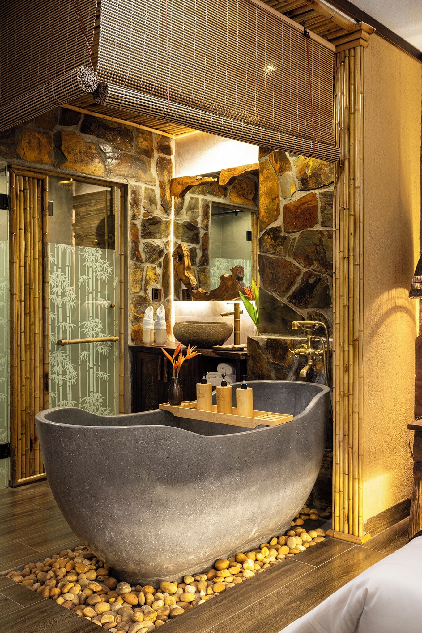 A stone-walled bathroom with a large bathtub, a wooden bench, and a bamboo shower area.