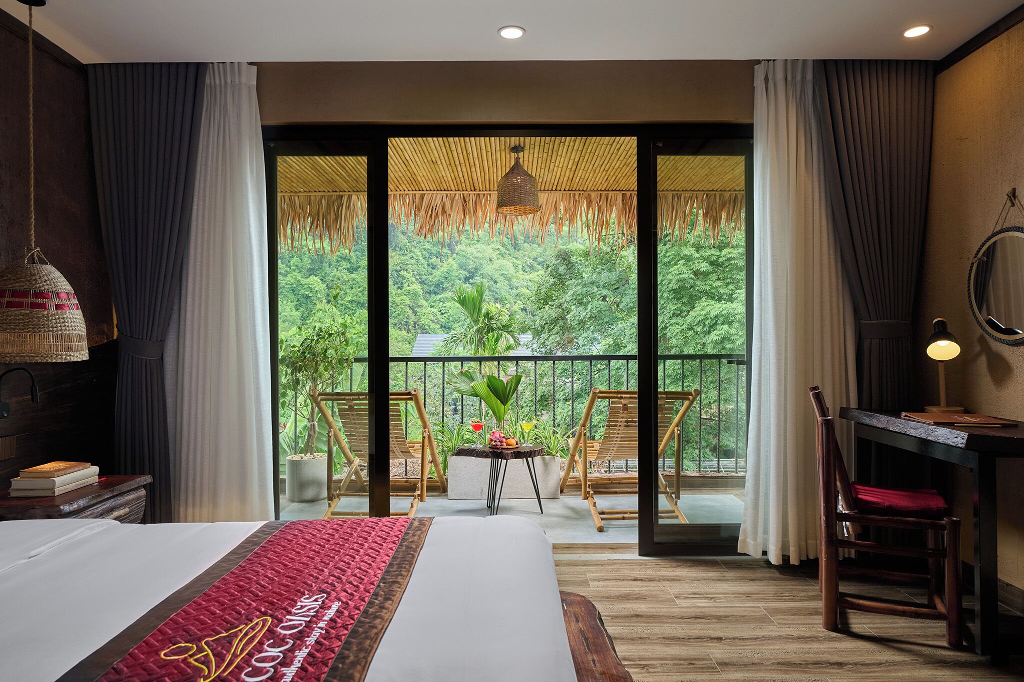 A hotel room with a large bed, a desk, a chair, a small table with plants, and a balcony with a view of greenery.