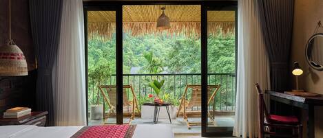 A hotel room with a large bed, a desk, a chair, a small table with plants, and a balcony with a view of greenery.