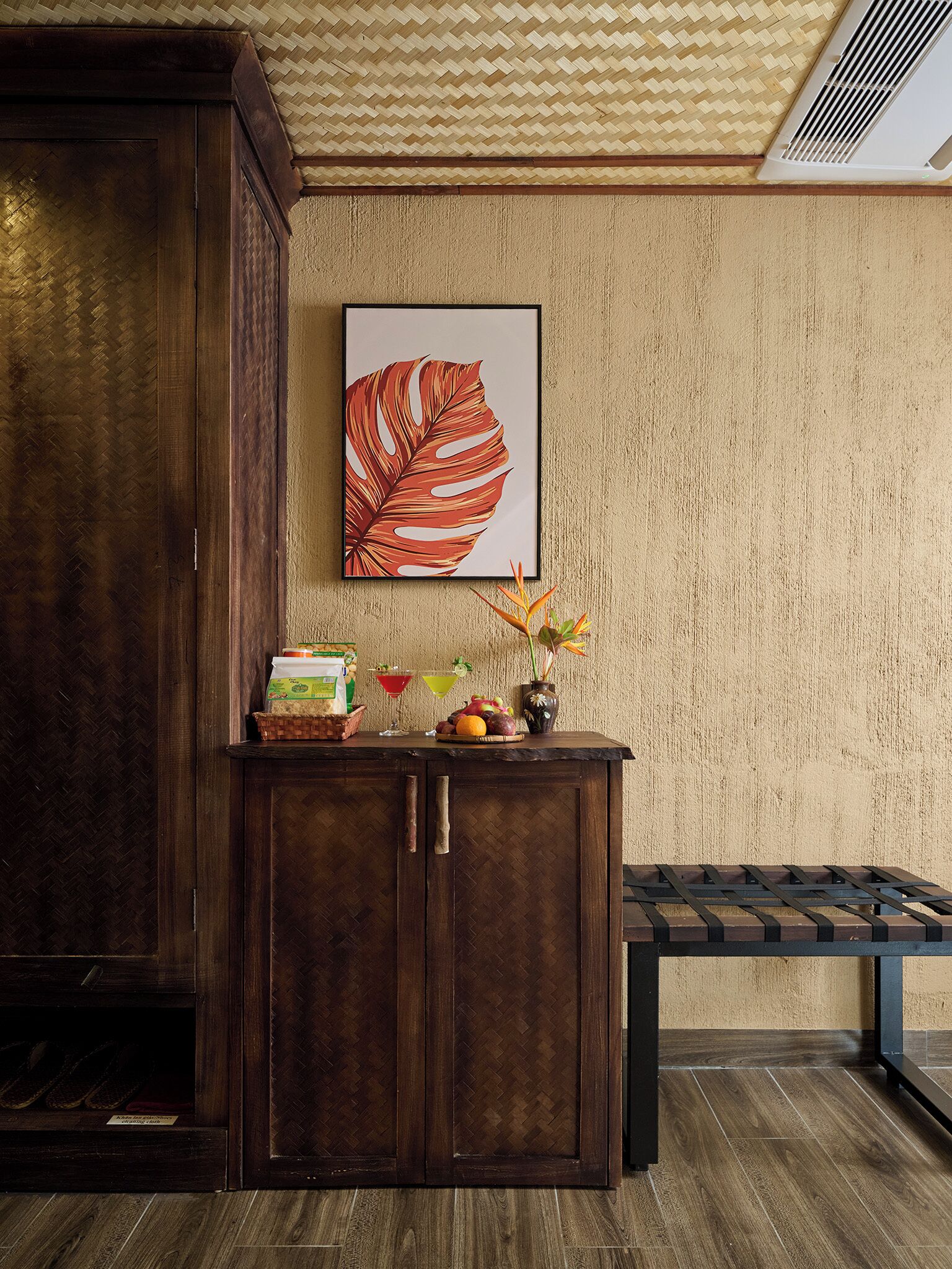 A wooden cabinet with a herringbone pattern, a framed tropical leaf artwork, and a small table with fruits and drinks.