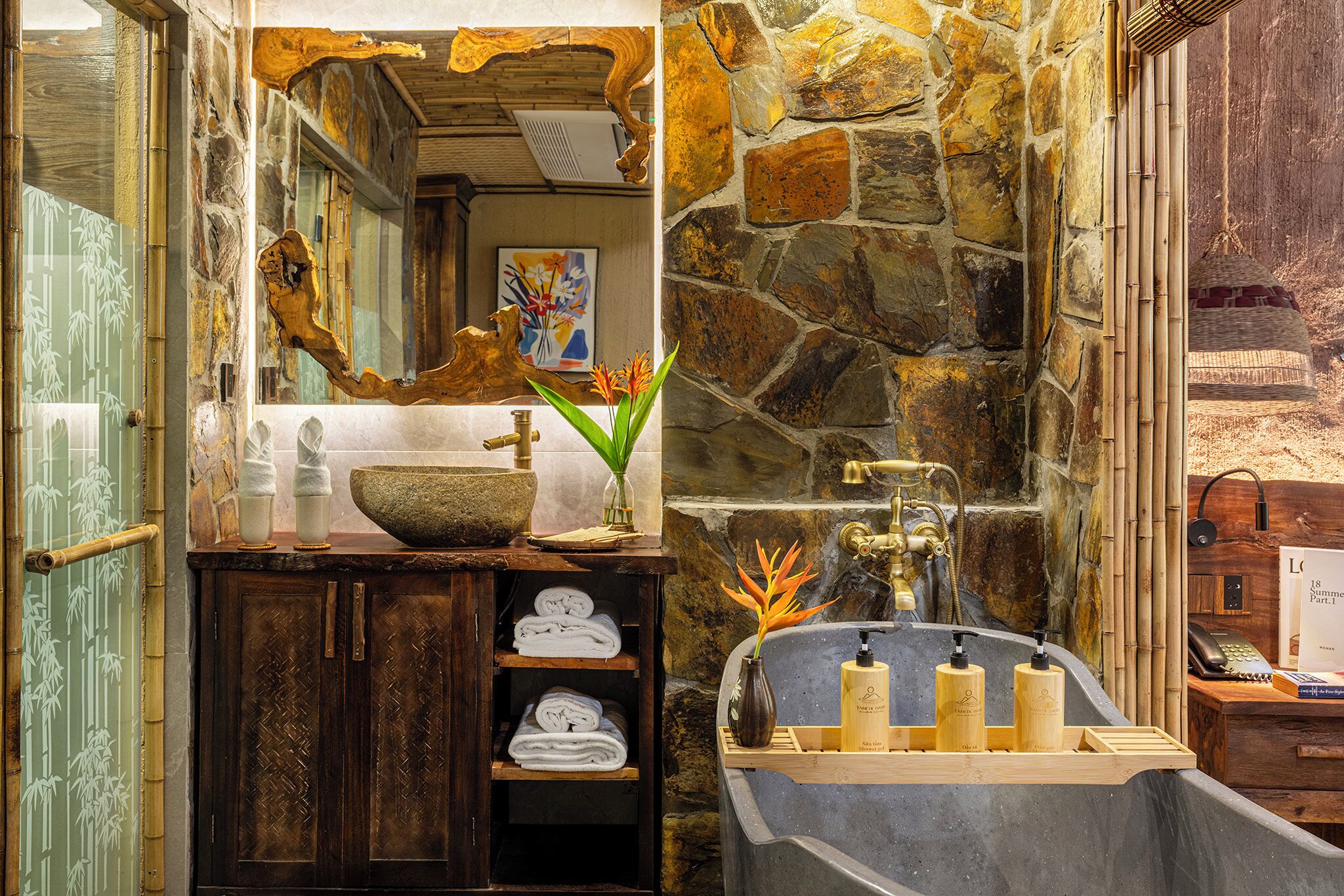 Une salle de bain avec un mur en pierre, un meuble-lavabo en bois, un grand miroir et une baignoire équipée de produits de toilette.