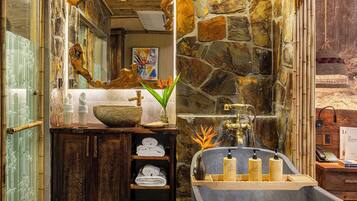 Une salle de bain avec un mur en pierre, un meuble-lavabo en bois, un grand miroir et une baignoire équipée de produits de toilette.