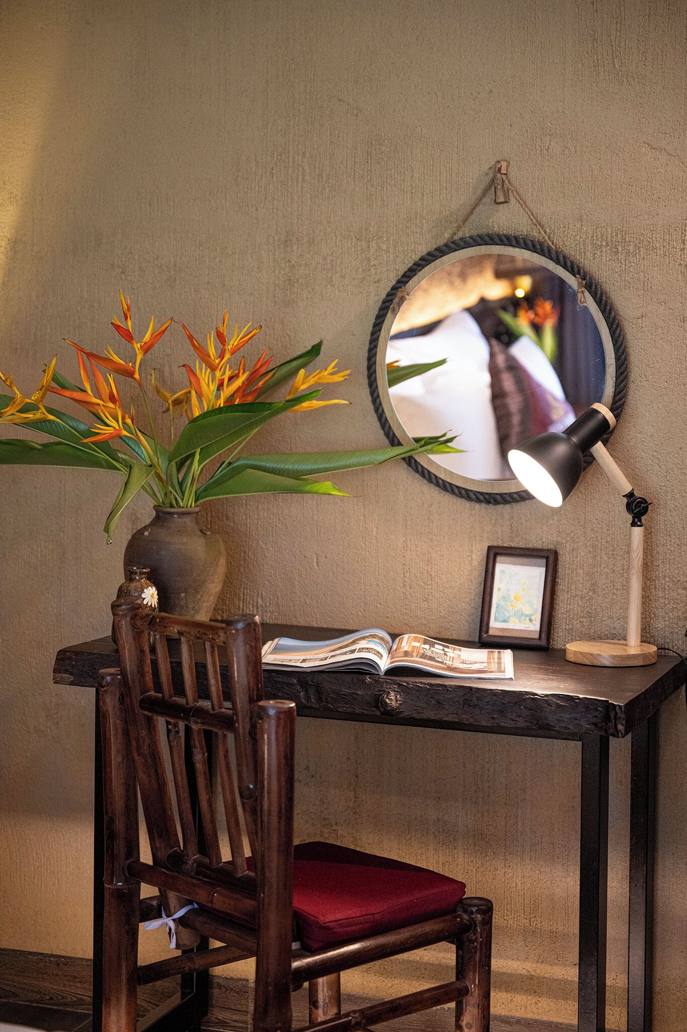 A wooden desk with a mirror, a lamp, a vase with flowers, and an open book.