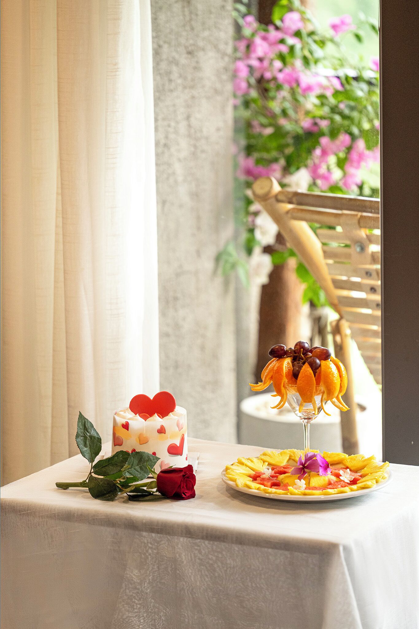 Une table avec un gâteau et une composition de fruits, une rose et une chaise avec un dossier en bois.