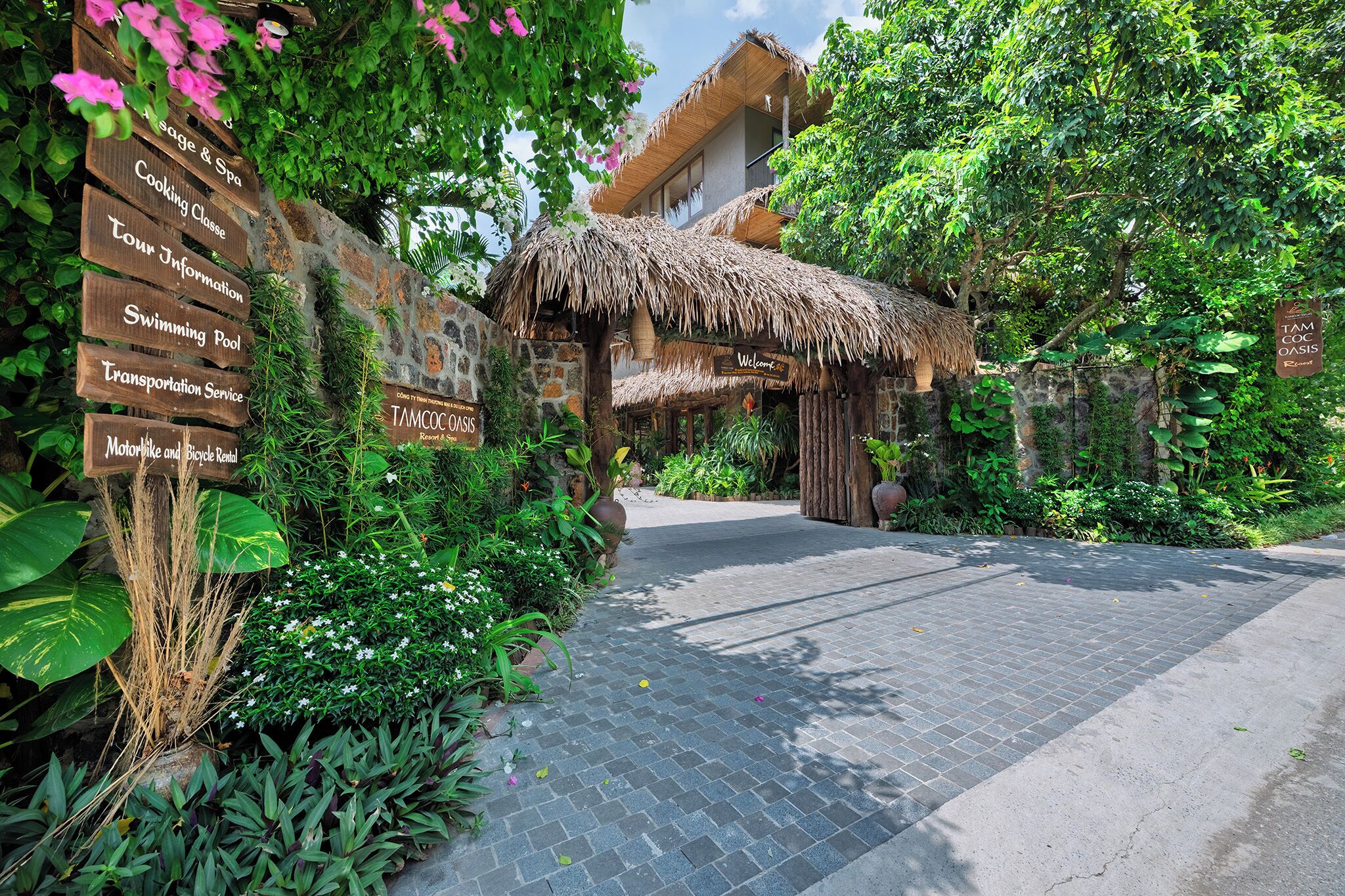 A tropical resort entrance with a thatched-roof structure, a signpost listing amenities like a spa, cooking class, and swimming pool, and a paved pathway leading to the building.