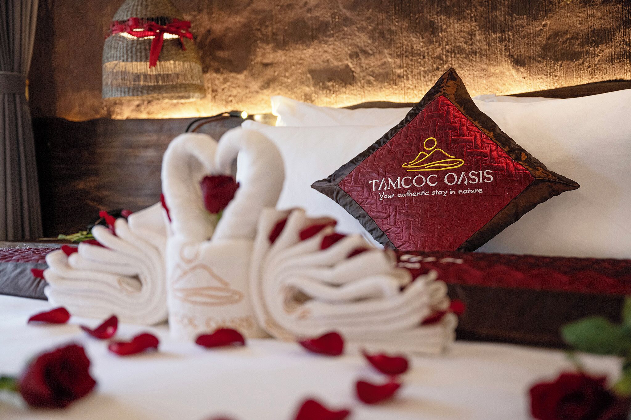 A neatly made bed with a swan-shaped towel arrangement, a pillow with 'Tamco Oasis' branding, and scattered rose petals.