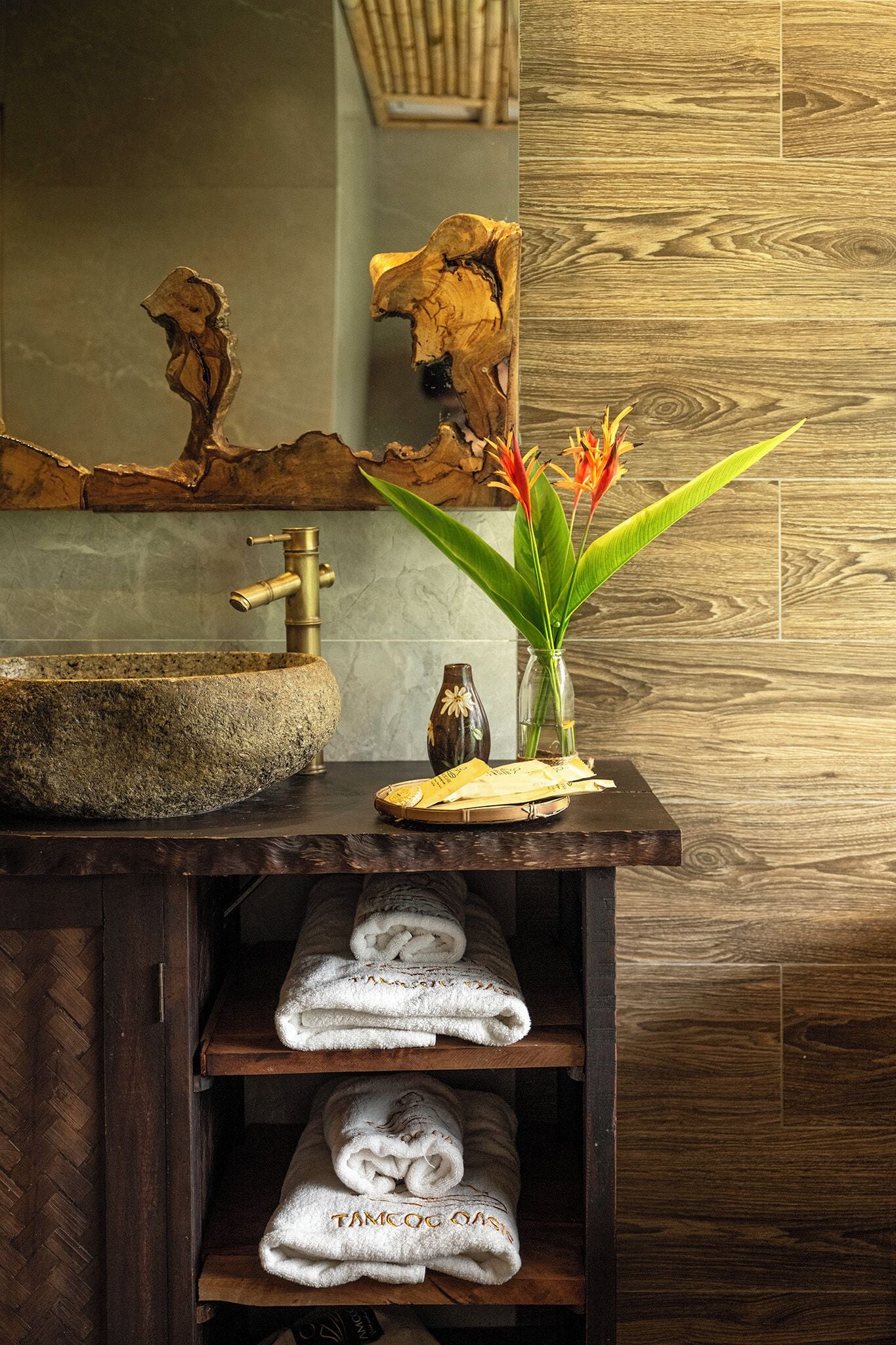 Un baño con lavamanos de piedra, tocador de madera, toallas y un jarrón con flores.