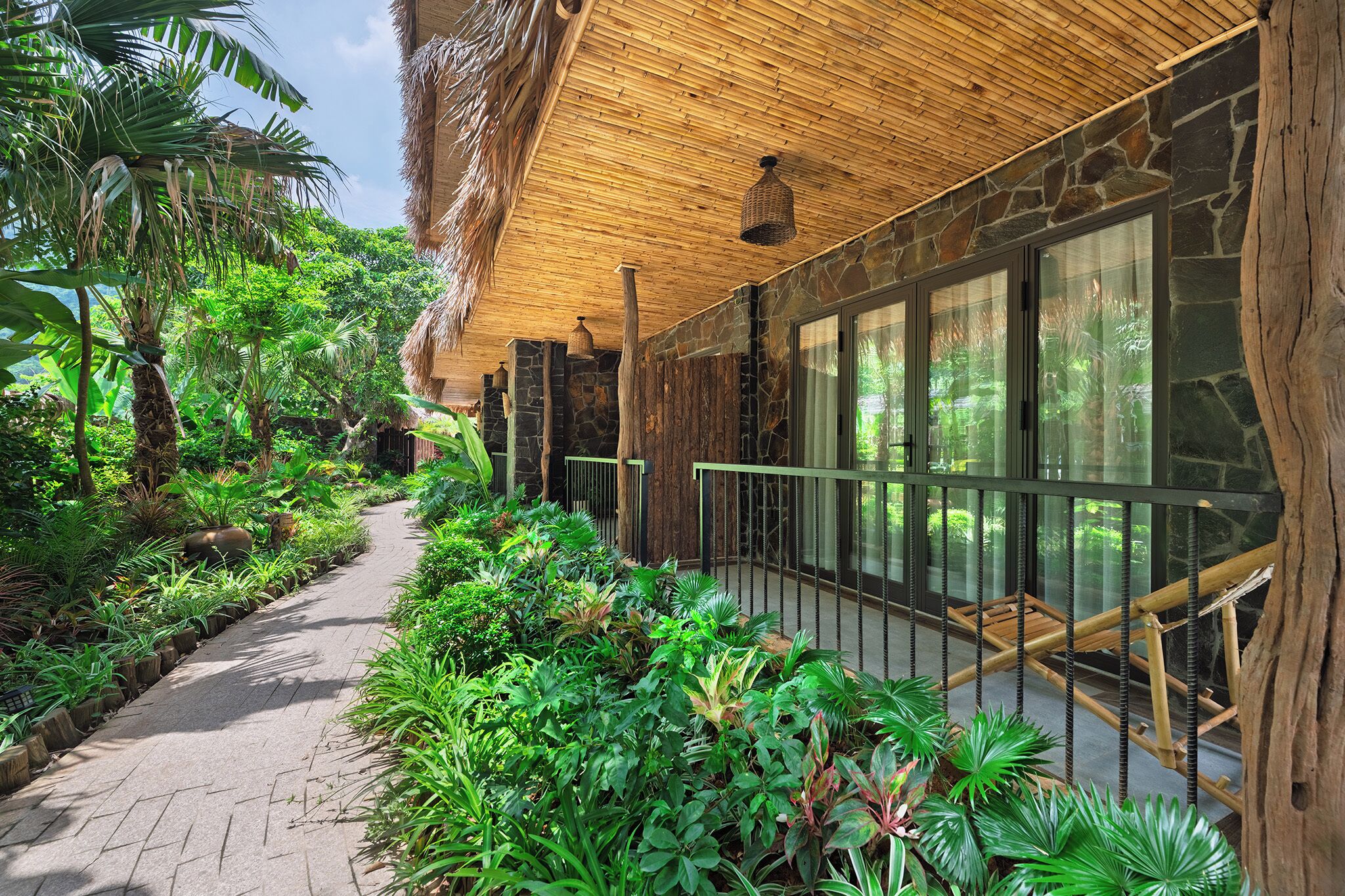 A pathway lined with lush greenery, a stone wall, and a wooden ceiling.