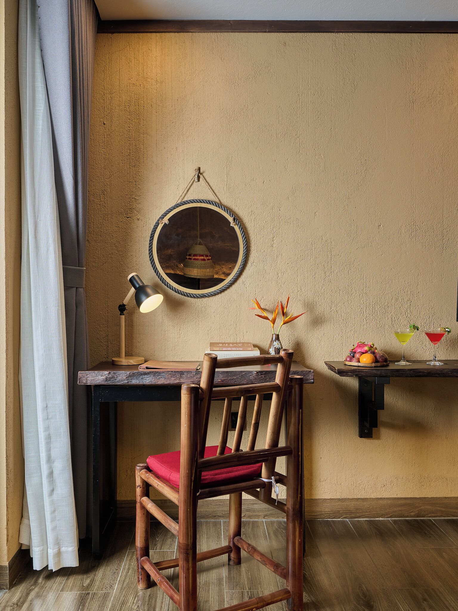 Un bureau en bois avec une lampe, une chaise, un miroir fixé au mur et une petite étagère avec des boissons et des fruits.