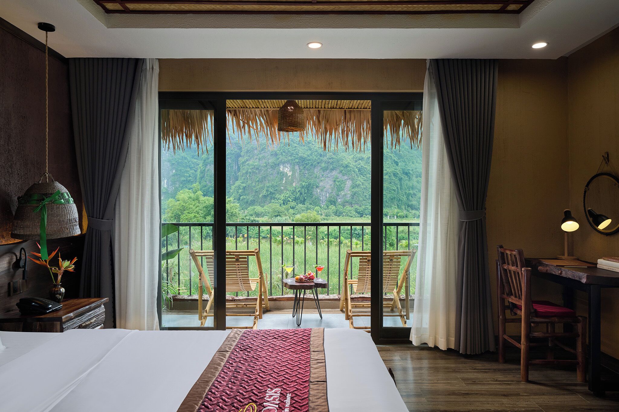 Habitación de hotel con una cama grande, un balcón con sillas, una mesita con una bebida y vistas a un área boscosa.