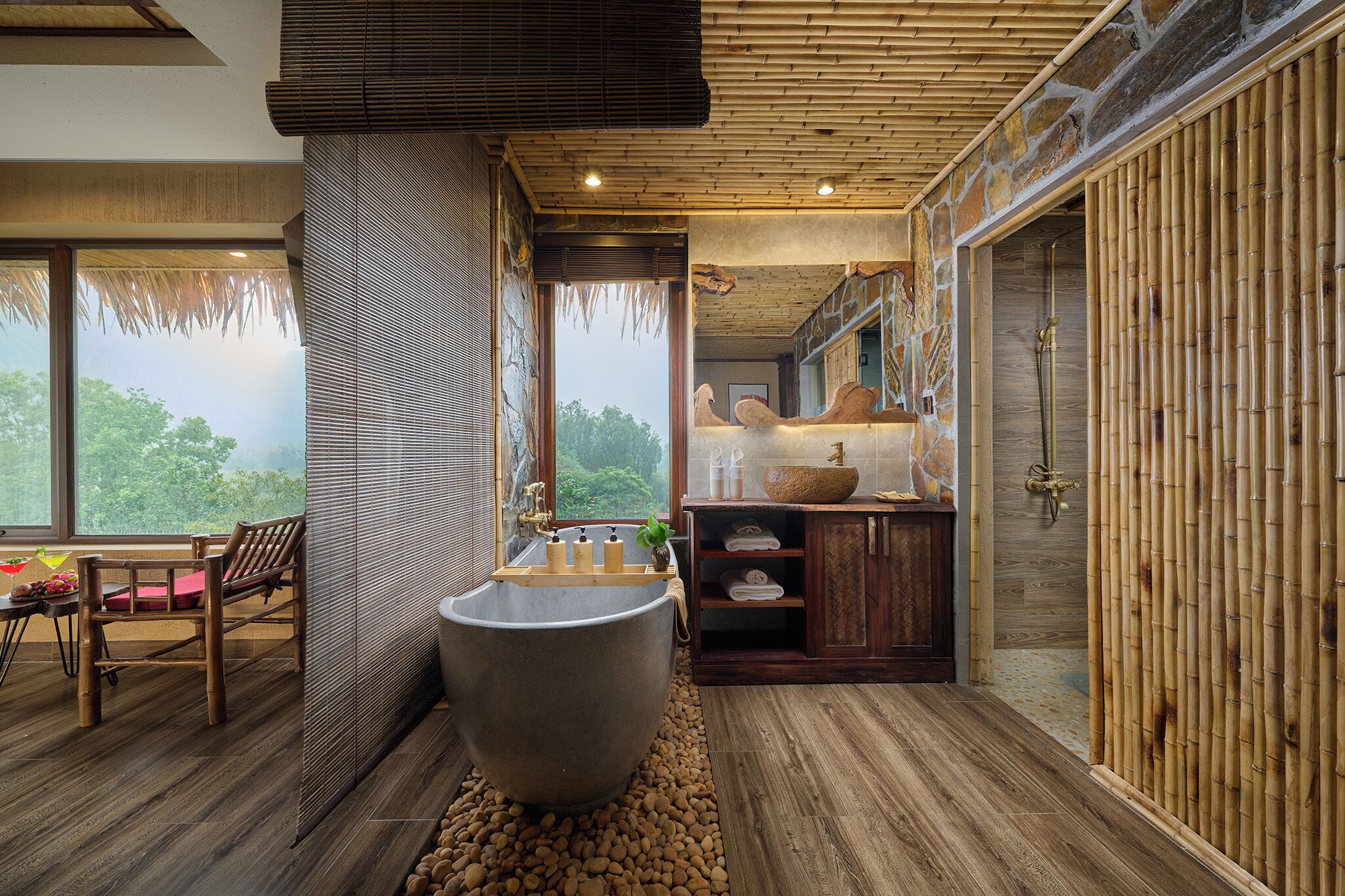 A bathroom with a large stone bathtub, wooden flooring, and a view of greenery outside.