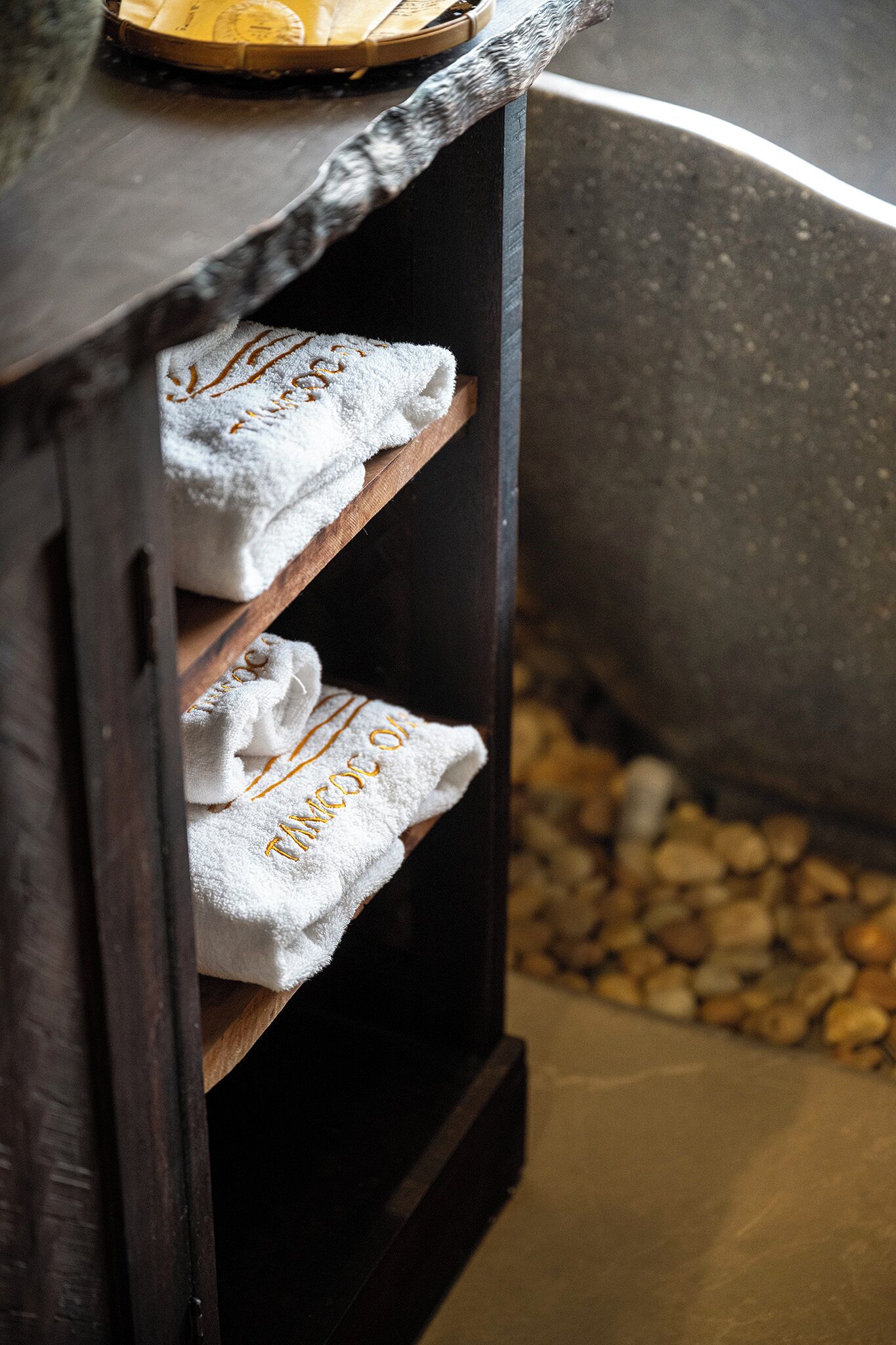 A wooden shelf with white towels, a bathtub, and a stone wall.