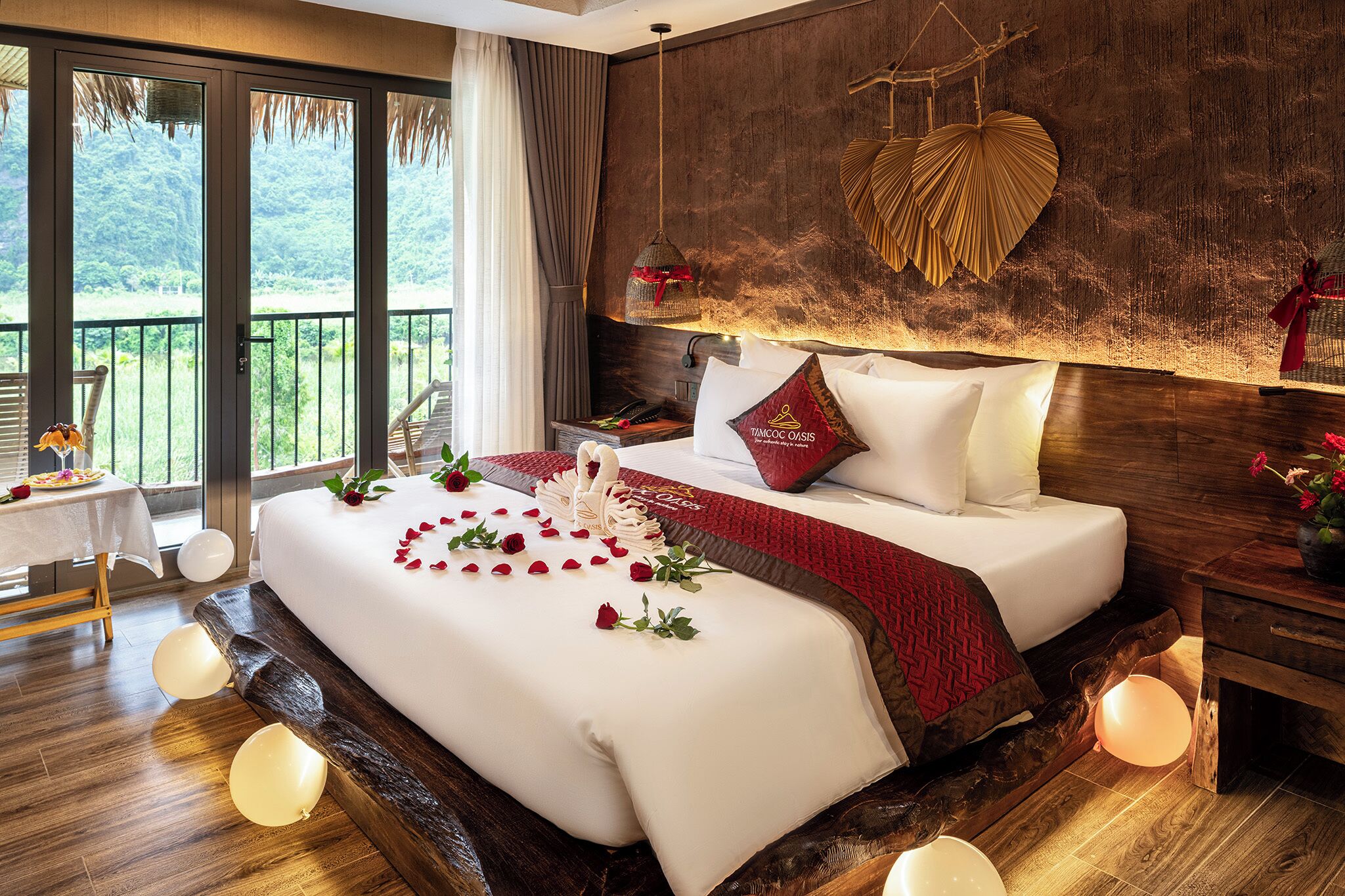 A hotel room with a large bed, a wooden headboard, a balcony with a view of greenery, and a table with a vase of flowers.