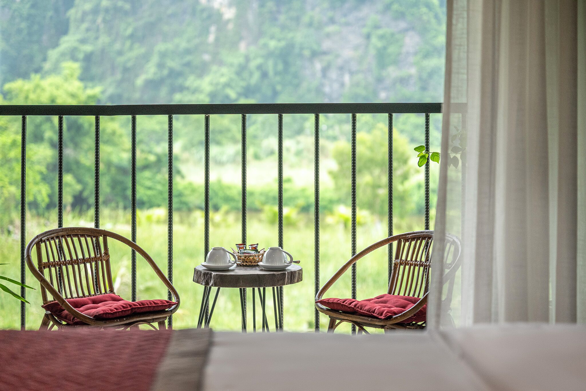 A balcony with wicker chairs, a small round table with cups and a teapot, and a view of a lush green landscape.