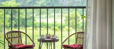 A balcony with wicker chairs, a small round table with cups and a teapot, and a view of a lush green landscape.