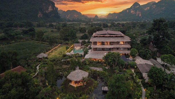 A large, multi-story building with a thatched roof, surrounded by lush greenery and a body of water. - Tam Coc Oasis Resort & Spa (Nam Hoa Lu)