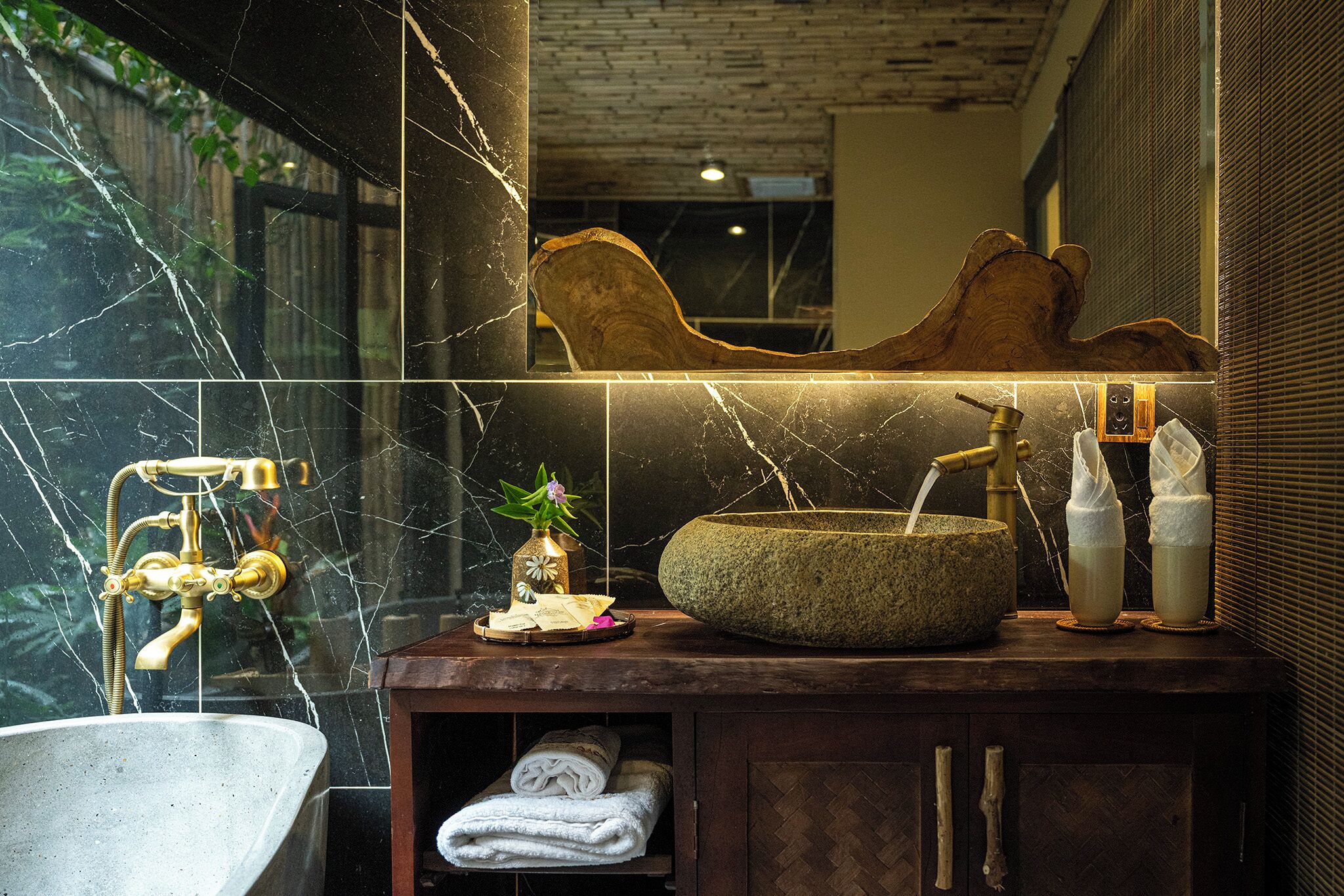 Un baño con lavamanos de mármol, un cuenco de madera, un perchero y un espejo que refleja una pared de ladrillo.