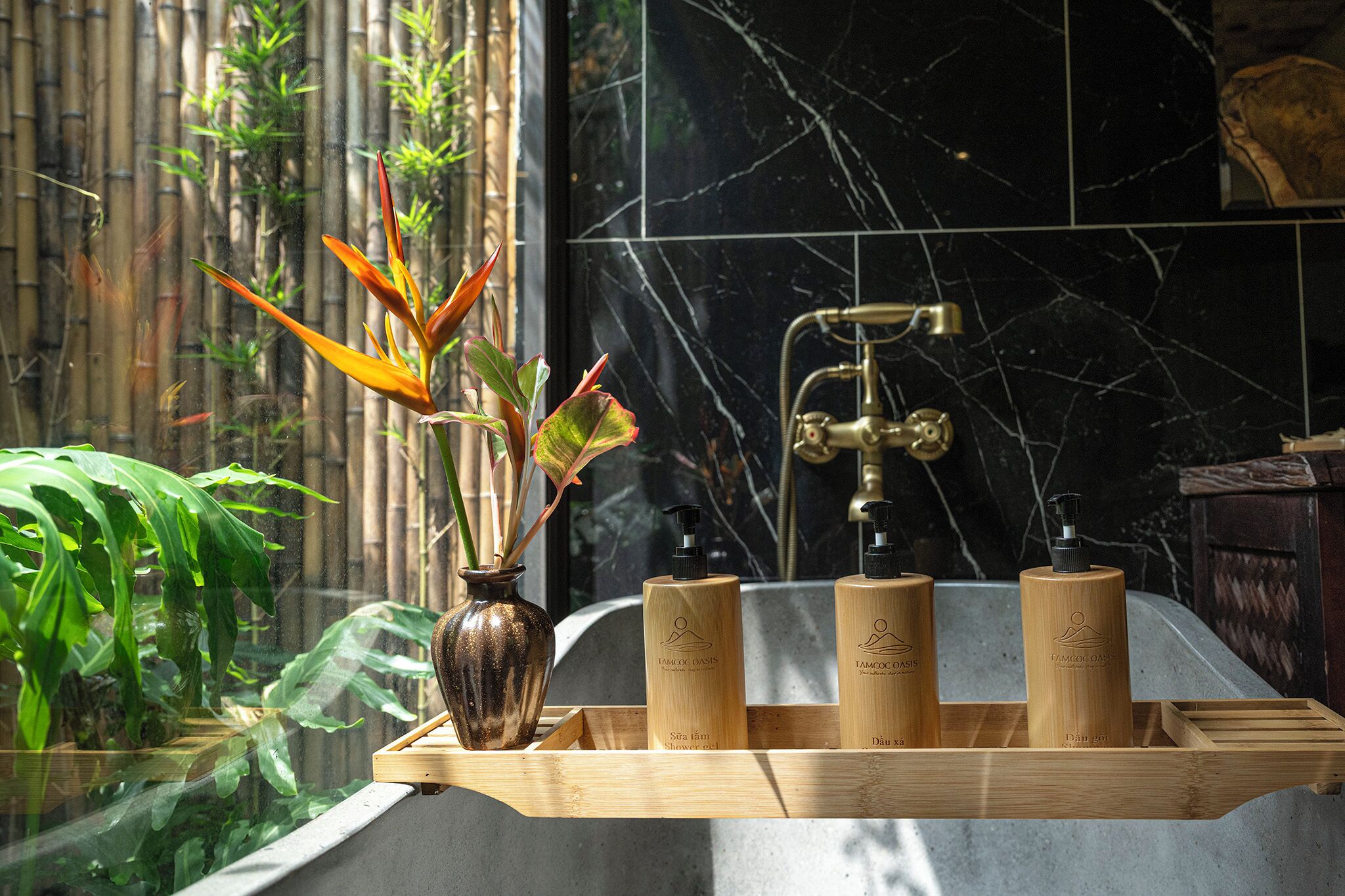 A bathroom with a wooden tray holding three bottles, a vase with plants, and a bathtub with a marble wall.