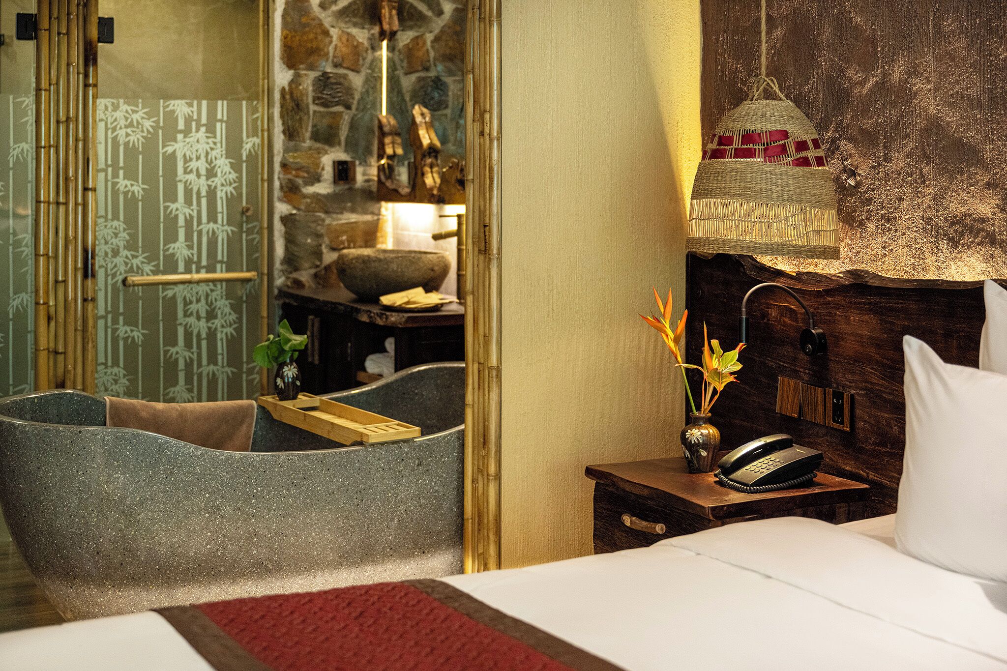 A hotel room with a stone wall, a bed with white bedding, a bedside table with a lamp, and a large window.