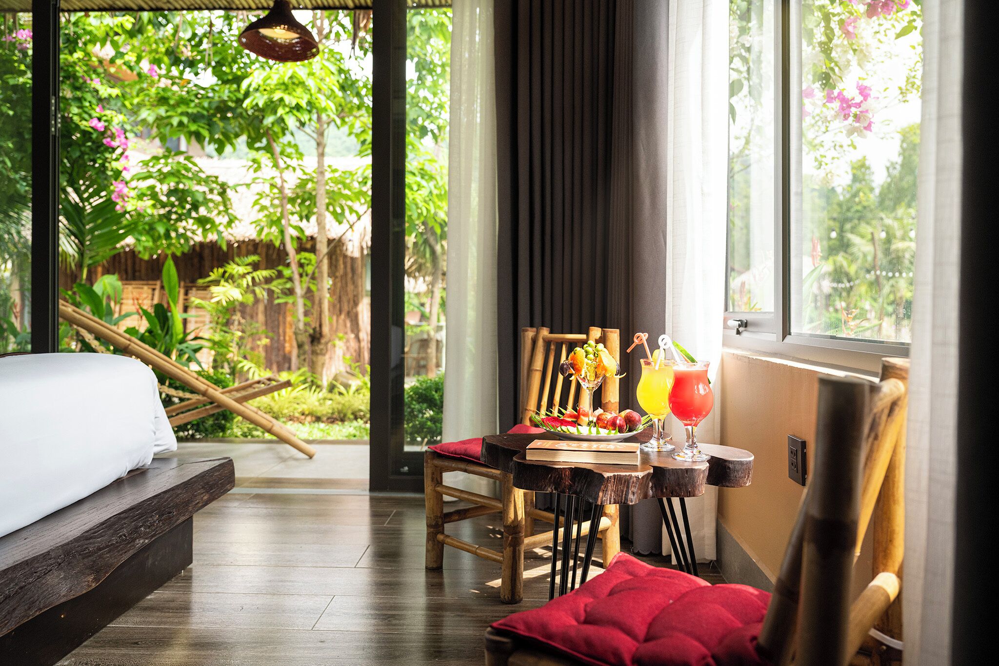 A room with a bed, a dining table with a fruit bowl and drinks, and a view of greenery outside.