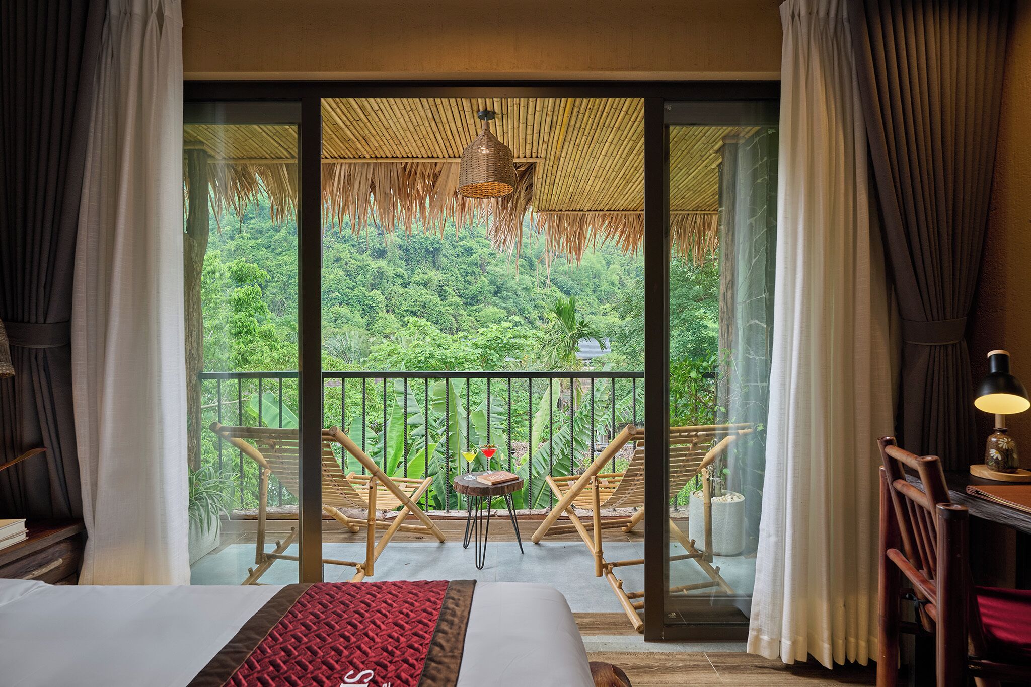 A room with a bed, a small table, two chairs, a lamp, and a view of lush greenery through a large window.