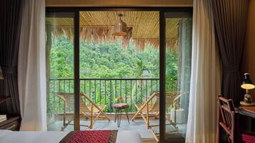 A room with a bed, a small table, two chairs, a lamp, and a view of lush greenery through a large window.