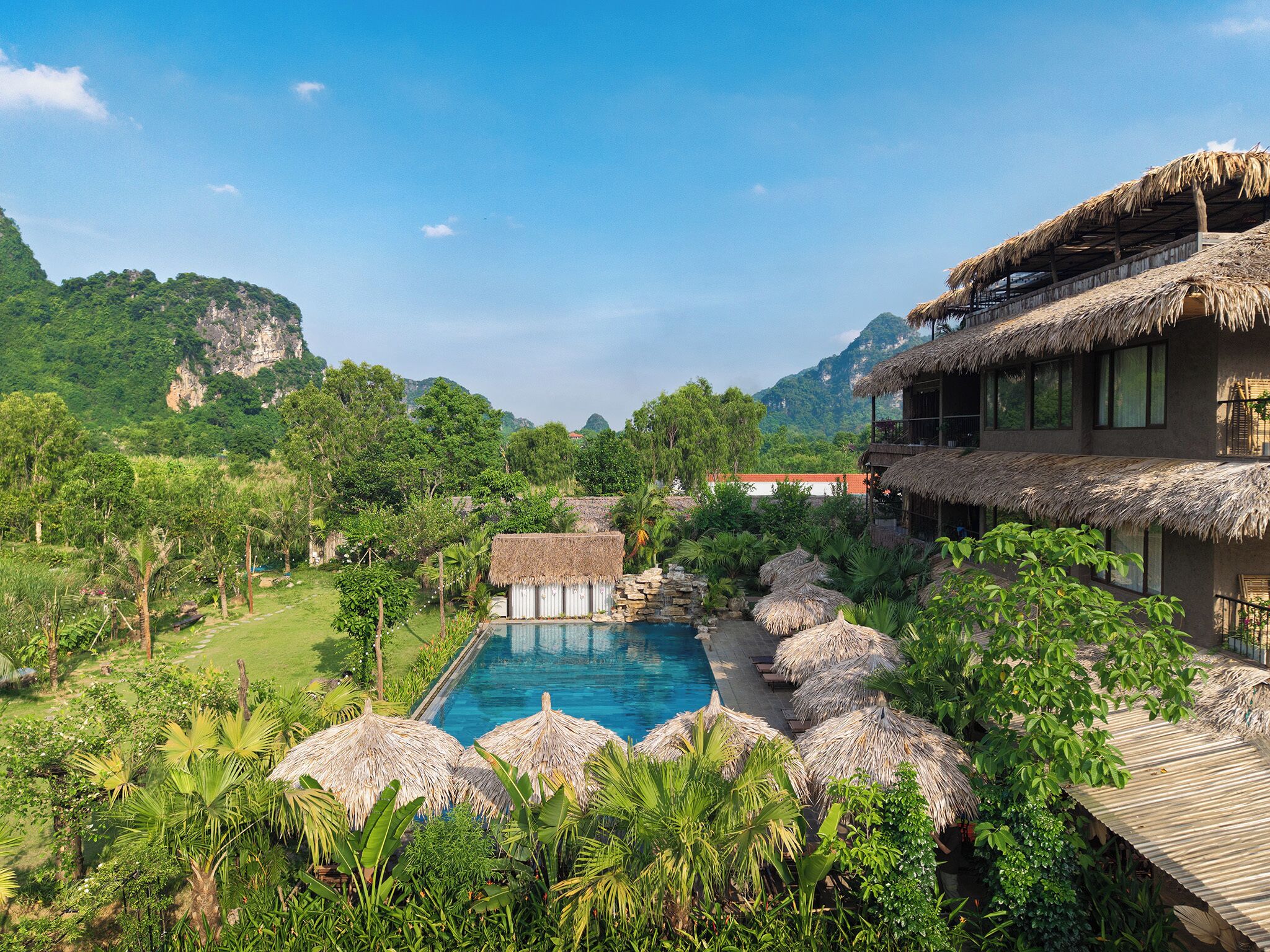 A resort with a pool, thatched-roof structures, lush greenery, and a mountainous backdrop.