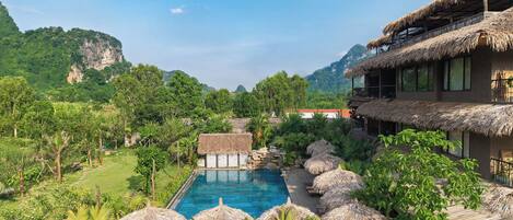 A resort with a pool, thatched-roof structures, lush greenery, and a mountainous backdrop.