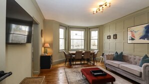 A living room with a sofa, a red ottoman, a dining table with chairs, and a TV mounted on the wall. - Grosvenor Abode, Royal Tunbridge Wells (Royal Tunbridge Wells)