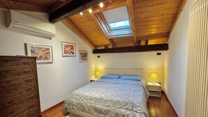 A bedroom with a sloped ceiling, wooden beams, a skylight, a bed with a patterned bedspread, two bedside tables with lamps, and a dresser. - Accogliente appartamento nel centro storico a 1 minuto dal mare (Sestri Levante)