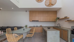 A modern kitchen with wooden cabinets, a central island, and a dining area with wicker chairs. - Villa Eudora 2br villa with private pool (Kec. Mengwi - Badung)