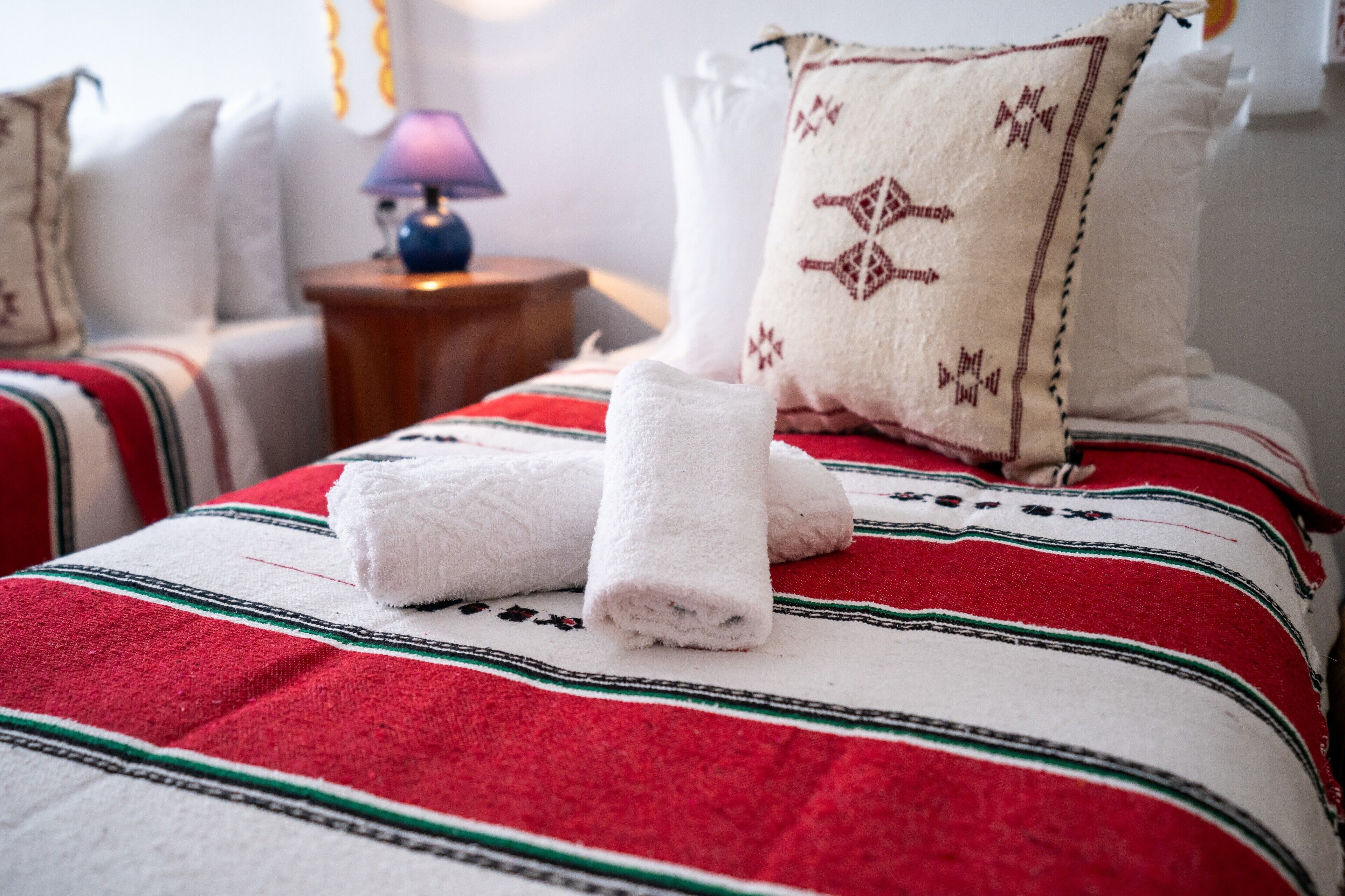 A neatly made bed with a red and white striped blanket, two rolled white towels, and a decorative pillow with a geometric pattern.