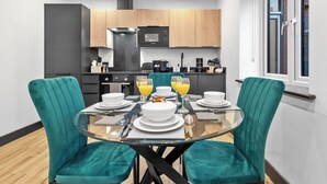 A modern kitchen with wooden cabinets, a black microwave, and a glass dining table set for four with teal chairs. - Luxury 1 Bedroom Flat with private patio Sleeps 4 (London)