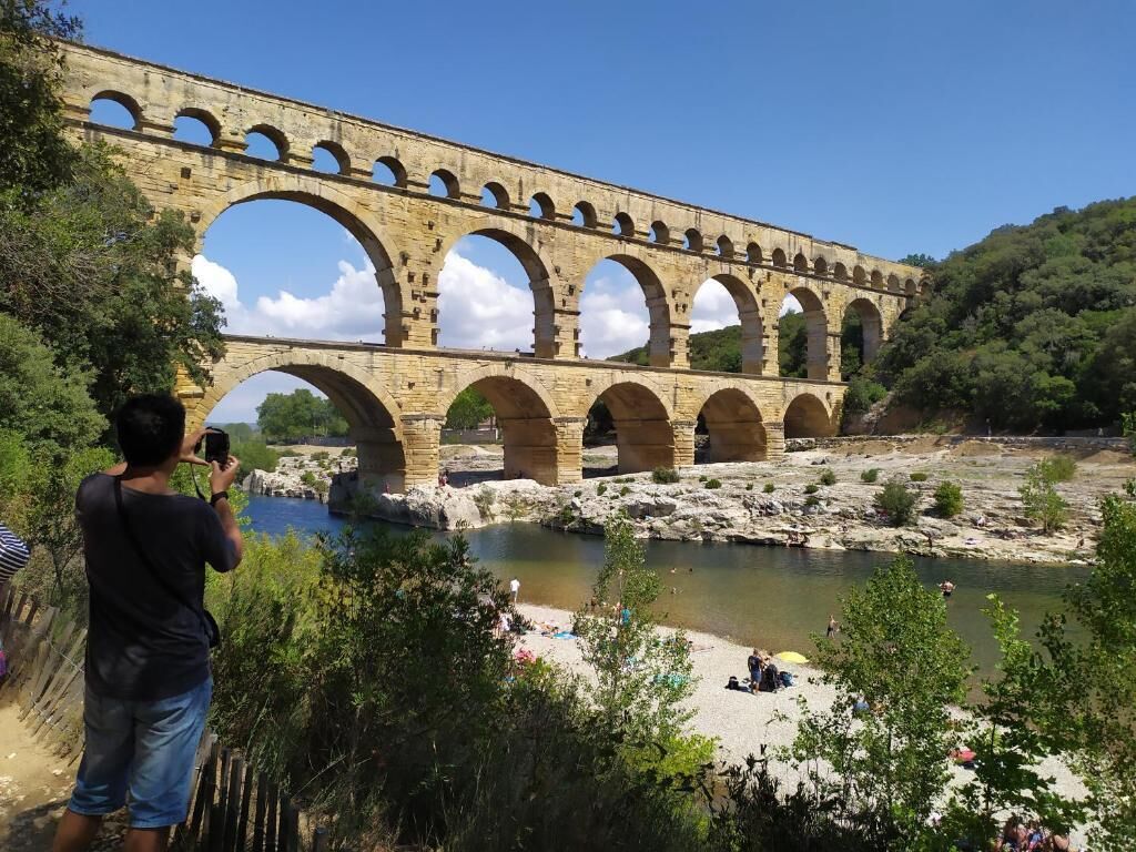 Ein aus Stein gefertigter Aquädukt mit mehreren Bögen überspannt einen Fluss, umgeben von viel Grün und blauem Himmel.