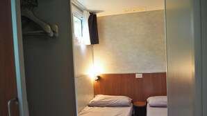 Quarto pequeno com duas camas, cabeceira de madeira, uma mesinha e uma janela com cortina.