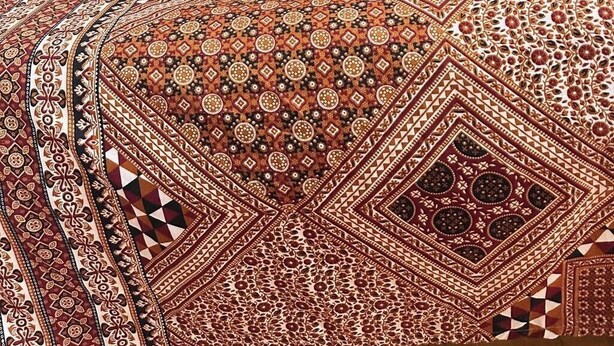 A neatly made bed with a patterned bedspread and a rolled white towel on the side.