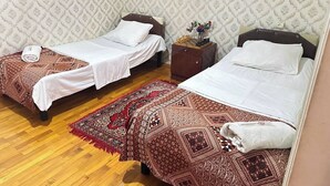 A room with two single beds, a wooden floor, patterned wallpaper, and a small nightstand with a vase of flowers.