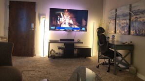 A living room with a flat-screen TV on a stand, a desk with a computer, and a chair. - Galaxy on Medical Mile Come Thrive! (Grand Rapids)