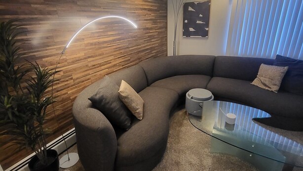 A modern living room with a curved grey sofa, a glass coffee table, and a wooden accent wall with recessed lighting. - Galaxy on Medical Mile Come Thrive! (Grand Rapids)