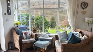 A cozy room with wicker furniture, a view of a green landscape, and a window with curtains. - Shantalla West Cork Farmhouse With Hot Tub & Sauna (Glanycarney)