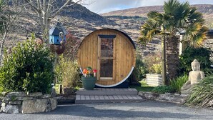 A wooden cabin with a circular door, surrounded by greenery and a mountain backdrop. - Shantalla West Cork Farmhouse With Hot Tub & Sauna (Glanycarney)