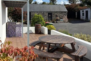 A wooden deck with a round table and benches, a stone building in the background, and a covered area with a metal roof.