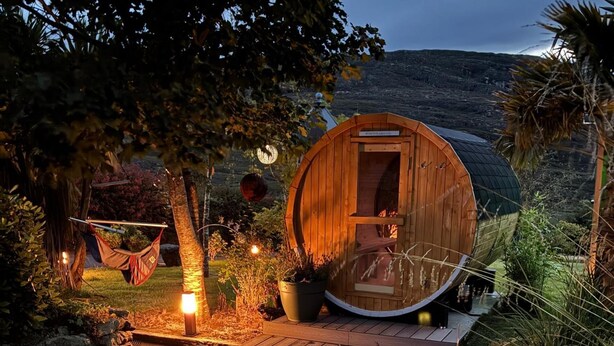 A wooden cabin with a door, surrounded by a garden with a hammock, potted plants, and a stone wall. - Shantalla West Cork Farmhouse With Hot Tub & Sauna (Glanycarney)