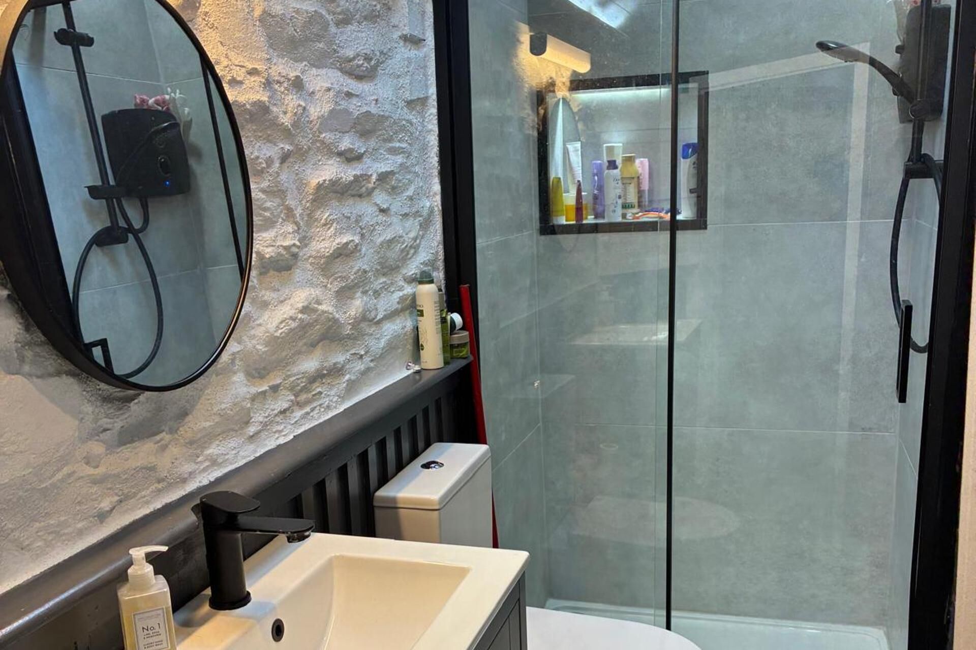 A modern bathroom with a stone wall, a large mirror, a sink with a soap dispenser, and a glass-enclosed shower area with toiletries on the wall.