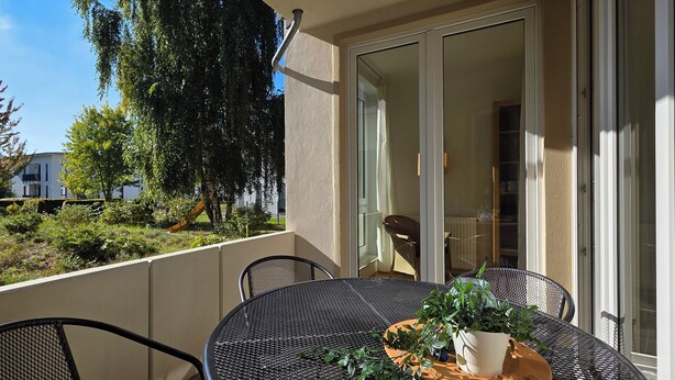A balcony with a table and chairs, a potted plant, and a view of a playground and trees. - Modern vacation apartment "Wellenkieker" with balcony and parking space in Graal-Müritz (Graal-Müritz)