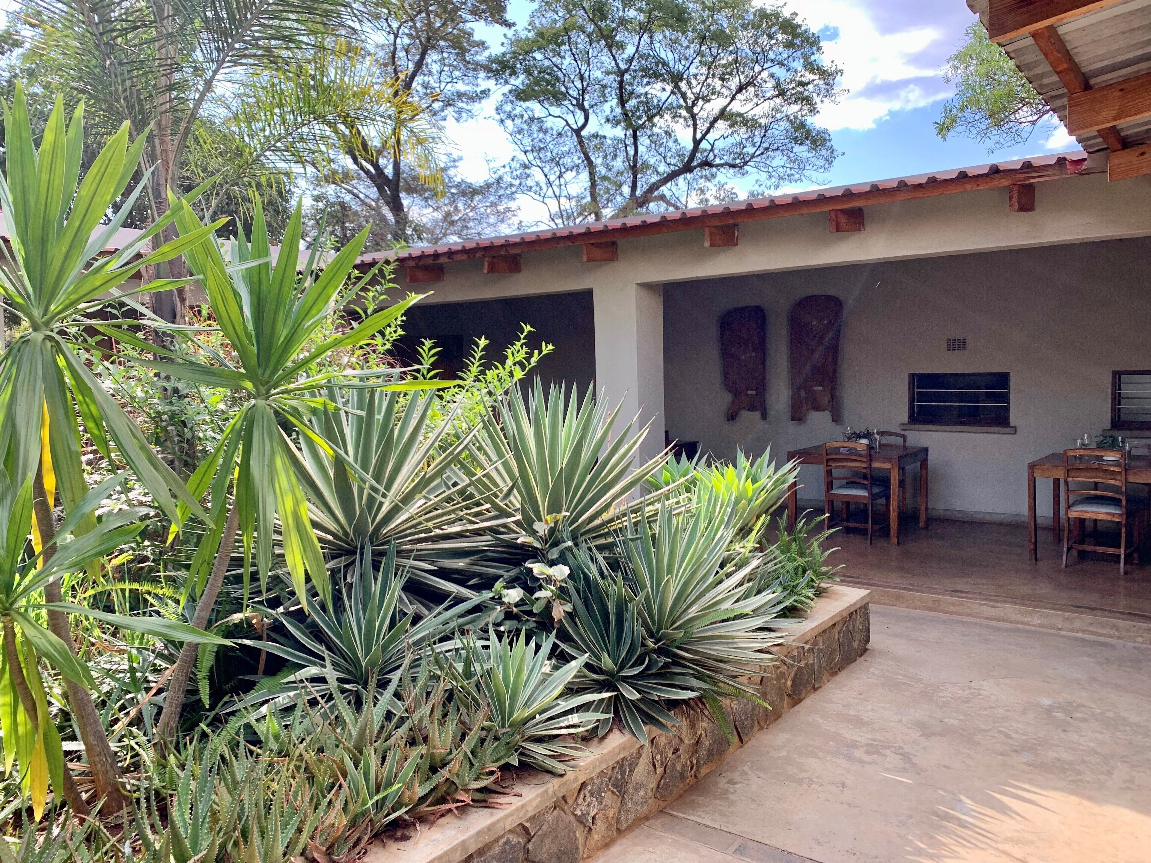 Un patio con una maceta de piedra llena de plantas, una mesa de madera con sillas, y un edificio con techo de tejas al fondo.