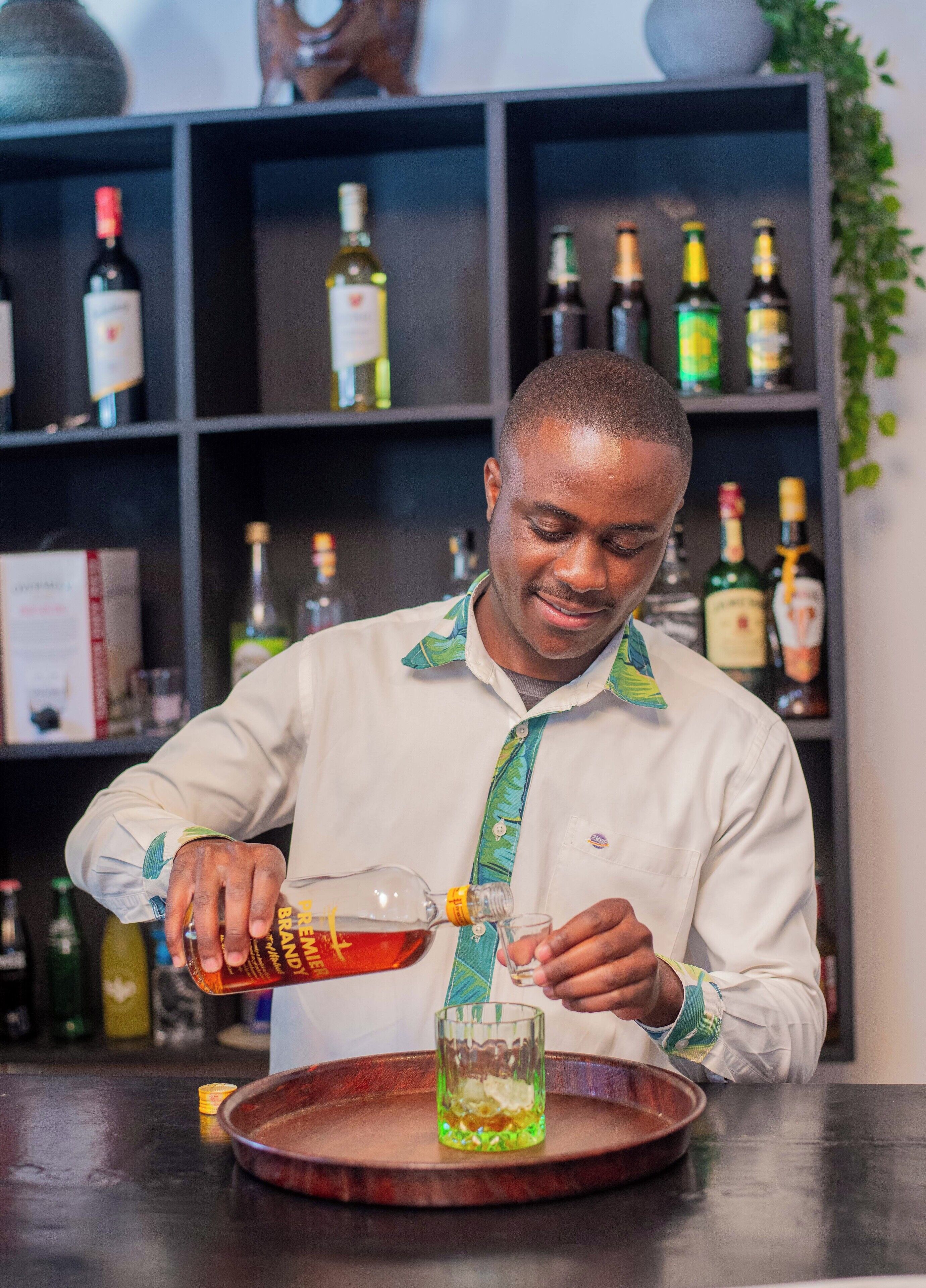 A person pouring a drink into a glass at a bar with shelves of bottles in the background.
