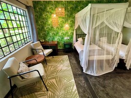 Una habitación con una cama bajo un dosel, una silla con cojín estampado, una mesita y una pared con follaje verde y luces colgantes.