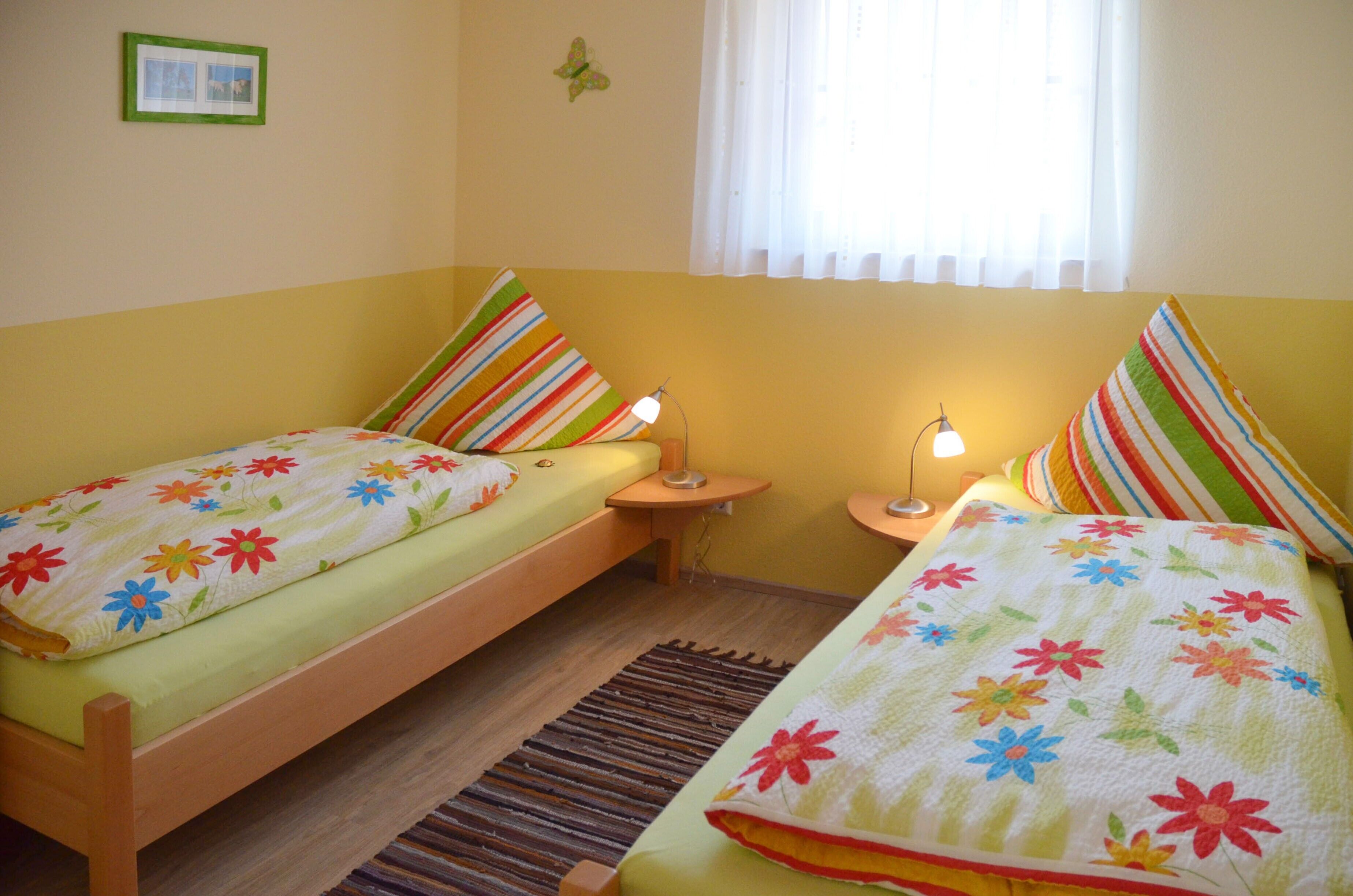 A small, cozy bedroom with two single beds, each with a colorful floral bedspread, a bedside table with a lamp, and a window with sheer curtains.