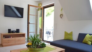 Ein Wohnzimmer mit einer Couch, einem Couchtisch mit Fernbedienung, einem an der Wand befestigten Fernseher, einer Schiebetür mit Glas und einem kleinen Holzschrank.